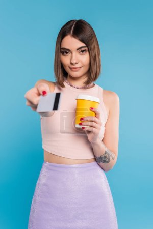 Photo for To go coffee, happy young woman with short hair, tattoos and nose piercing holding paper cup and giving credit card on blue background, generation z, summer trends, attractive, coffee culture - Royalty Free Image
