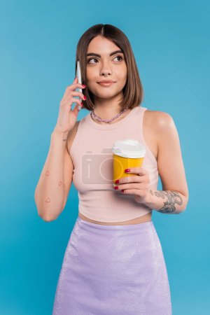to go coffee, happy and tattooed young woman with short hair and nose piercing holding paper cup and talking on smartphone on blue background, generation z, summer trends, attractive, coffee culture 