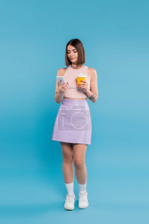 to go coffee, tattooed young woman with short hair and nose piercing holding paper cup and talking on smartphone on blue background, generation z, summer trends, attractive, full length