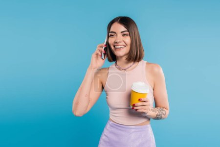 to go coffee, joyful and tattooed young woman with short hair and nose piercing holding paper cup and talking on smartphone on blue background, generation z, attractive, coffee culture 