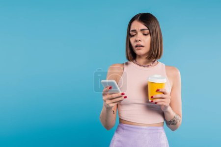to go coffee, social media influencers, tattooed young woman with short hair and nose piercing holding paper cup and using smartphone on blue background, generation z, summer trends 