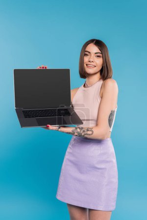 Photo for Freelancer, cheerful young woman with short hair, tattoos and nose piercing holding laptop on blue background, generation z, summer trends, attractive, remote work, everyday style, blank screen - Royalty Free Image