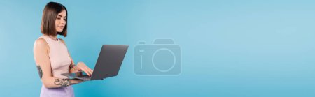 Freiberuflerin, brünette junge Frau mit kurzen Haaren, Tätowierungen und Nasenpiercing mit Laptop auf blauem Hintergrund, Generation Z, Sommertrends, attraktiv, Fernarbeit, Alltagsstil, Banner 