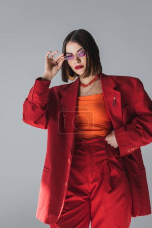 lady in red, young brunette woman with short hair posing in pink sunglasses and red suit on grey background, generation z, trendy outfit, fashion model, professional attire 