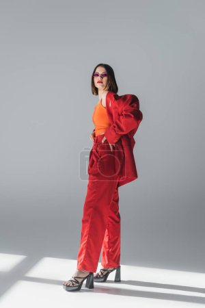 business fashion, young brunette woman with short hair posing in pink sunglasses and red suit on grey background, generation z, trendy outfit, fashionable model, full length 