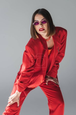 red outfit, young brunette woman with short hair posing in pink sunglasses and suit on grey background, generation z, trendy outfit, fashionable model, professional attire, executive style 