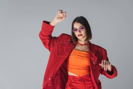 fashionable gen z, young brunette woman with short hair posing in pink sunglasses and red suit on grey background, generation z, trendy outfit, fashion model, professional attire 