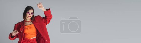 Foto de Moda gen z, joven morena con el pelo corto posando en gafas de sol de color rosa y traje rojo sobre fondo gris, generación z, traje de moda, modelo de moda, estilo ejecutivo, bandera - Imagen libre de derechos