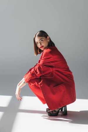 full length, lady in red, young brunette woman with short hair sitting in suit on grey background, generation z, trendy outfit, fashionable model, professional attire, executive style, heeled shoes