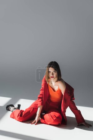 volle Länge, junge Frau mit kurzen Haaren im Anzug sitzend auf grauem Hintergrund, Generation Z, trendiges Outfit, modisches Modell, professionelle Kleidung, Führungsstil, hohe Schuhe, Dame in Rot 