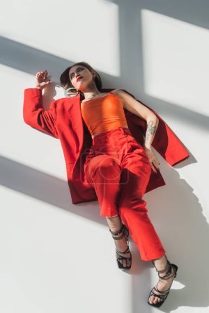 vue de dessus, jeune femme tatouée avec les cheveux courts couché en costume rouge sur fond gris, génération z, modèle à la mode, tenue professionnelle, mode d'entreprise, chaussures à talons, dame en rouge 