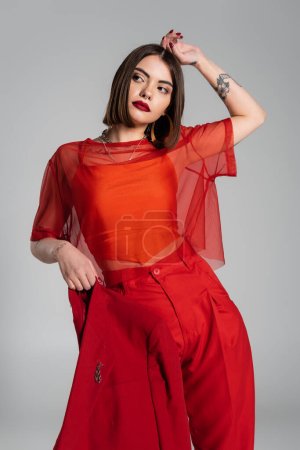 maquillaje audaz, moda corporativa, mujer joven tatuada con pelo corto sosteniendo chaqueta roja sobre fondo gris, generación z, traje de moda, modelo de moda, atuendo profesional 