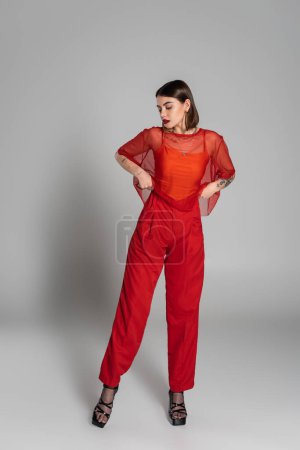 red outfit, tattooed and brunette woman with short hair and nose piercing posing in transparent blouse and pants on grey background, modern style, generation z, fashion photography, full length 