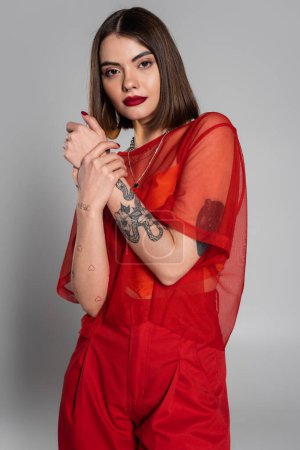 fashionista, traje rojo, tatuado y mujer joven con pelo corto y piercing en la nariz posando en blusa transparente y pantalones sobre fondo gris, estilo moderno, generación z, moda