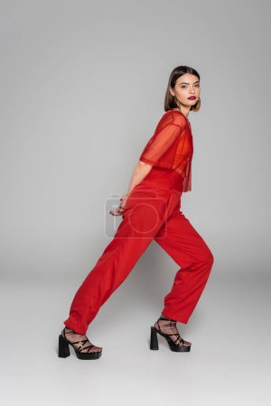model in red outfit, tattooed and brunette woman with short hair and nose piercing posing in transparent blouse and pants on grey background, modern style, generation z, fashion trend, full length 