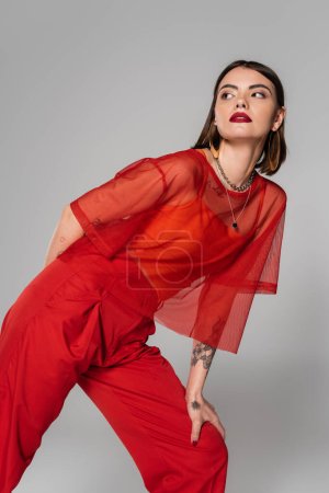 modelo en traje rojo, mujer joven tatuada con pelo corto y piercing en la nariz posando en blusa transparente y pantalones sobre fondo gris, estilo moderno, generación z, tendencia de la moda 