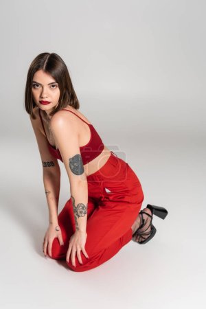 young model in red outfit, tattooed brunette woman with short hair and nose piercing posing in red crop top and pants on grey background, modern style, generation z, fashion trend 