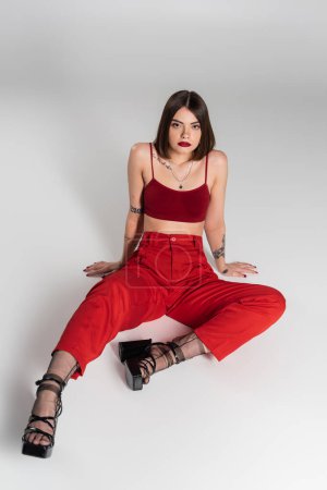 fashion trend, young model in red outfit, tattooed woman with short hair and nose piercing posing in red crop top and pants on grey background, generation z, full length, high angle view 