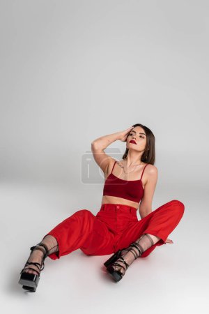 fashion trend, chic style, young model in red outfit, tattooed woman with short hair and nose piercing posing in red crop top and pants on grey background, generation z, full length 