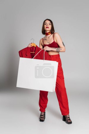 consumerism, young tattooed woman with short hair and nose piercing holding hanger with blazer and shopping bag on grey background, modern fashion trend, chic style, neck scarf, full length 