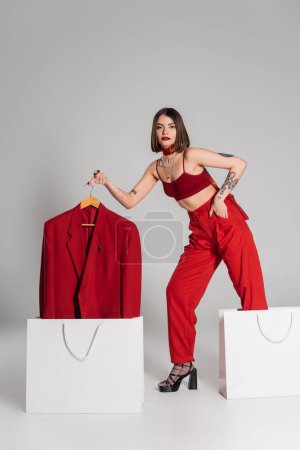 consumerism, tattooed woman with short hair and nose piercing holding hanger with blazer and taking it out of shopping bag on grey background, modern fashion trend, hand in pocket, full length 