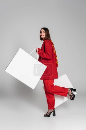 consumerism, happy woman with brunette short hair and nose piercing holding shopping bags and walking on grey background, modern fashion trend, fashionable outfit, red suit, full length 