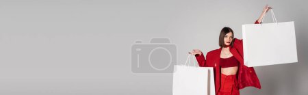 consumerism, young woman with brunette short hair and nose piercing holding shopping bags and walking on grey background, modern fashion trend, fashionable outfit, red suit, banner 