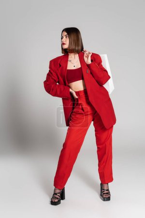 roter Anzug, fettes Make-up, tätowierte junge Frau mit brünetten kurzen Haaren und Nasenpiercing, Einkaufstaschen haltend und auf grauem Hintergrund stehend, Jugendkultur, modisches Outfit, Konsum 