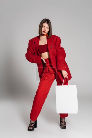 red suit, stylish posing, tattooed young woman with brunette short hair and nose piercing holding shopping bags and standing on grey background, youth culture, fashionable outfit, consumerism 