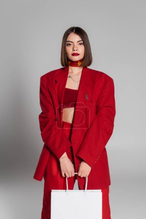 fashionable, bold makeup, young woman with brunette short hair and nose piercing holding shopping bags and standing on grey background, youth culture, trendy outfit, red suit, consumerism