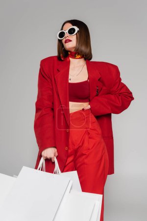 moda, generación z, mujer joven con pelo corto morena y piercing en la nariz posando en gafas de sol y sosteniendo bolsas de compras sobre fondo gris, dama de rojo, consumismo 