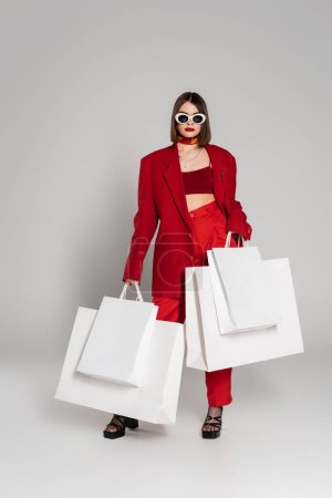 Generation z, junge Frau mit brünetten kurzen Haaren und Nasenpiercing posiert in Sonnenbrille und rotem Anzug, während sie Einkaufstüten auf grauem Hintergrund hält, moderne Mode, Konsum, volle Länge  