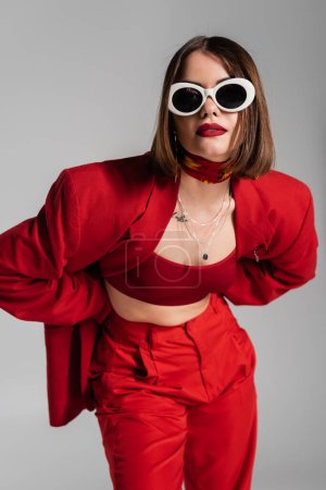 Generation Z, tätowierte junge Frau mit brünetten kurzen Haaren und Nasenpiercing posiert in Sonnenbrille und rotem Anzug auf grauem Hintergrund, moderne Mode, trendiges Outfit, schicker Stil 
