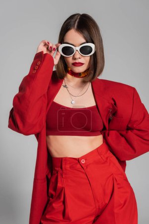 Trendiges Outfit, Generation Z, tätowierte junge Frau mit brünetten kurzen Haaren und Nasenpiercing posiert in Sonnenbrille und rotem Anzug auf grauem Hintergrund, moderne Mode, schicker Stil 