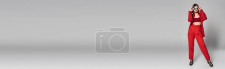 Foto de Generación z, mujer joven tatuada con pelo corto y piercing en la nariz posando en gafas de sol y traje rojo sobre fondo gris, moda moderna, atuendo de moda, estilo elegante, longitud completa, pancarta - Imagen libre de derechos
