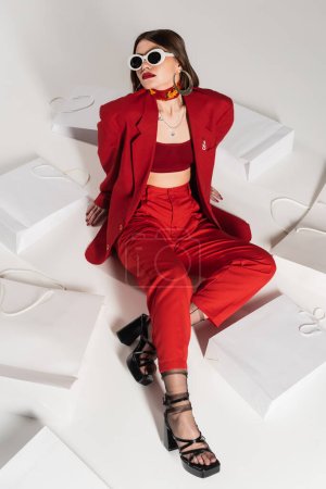 consumerism, young woman with brunette short hair and nose piercing and tattoo posing in sunglasses and red suit while sitting around shopping bags on grey background, high angle view, full length