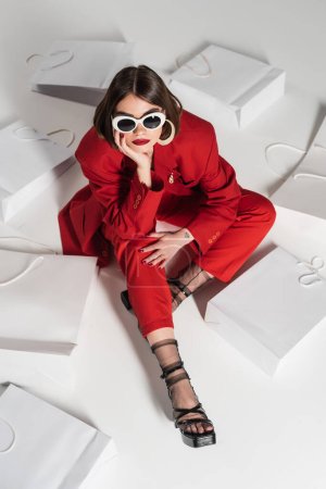 consumismo, mujer joven con pelo corto morena, piercing en la nariz y tatuaje posando en gafas de sol y traje rojo mientras se sienta alrededor de bolsas de compras sobre fondo gris, vista de ángulo alto 