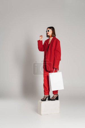 génération z, consumérisme, jeune femme aux cheveux courts bruns et nez percé posant en lunettes de soleil et costume rouge tout en tenant un sac à provisions et debout sur un cube de béton sur fond gris 