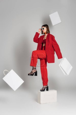 generation z, consumerism, fashion model with brunette short hair and nose piercing posing in sunglasses and red suit while standing on concrete cube around flying shopping bags on grey background 