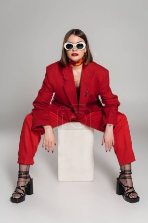 Foto de Generación z, modelo de moda con pelo corto morena y piercing en la nariz posando en gafas de sol y traje rojo mientras está sentado en cubo de hormigón sobre fondo gris, señora de rojo, fashionista - Imagen libre de derechos