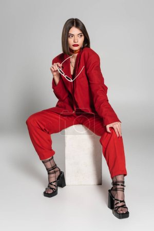 Photo for Fashion model with brunette short hair and nose piercing posing in red suit while holding sunglasses and sitting on concrete cube on grey background, lady in red, young woman, fashion trend - Royalty Free Image
