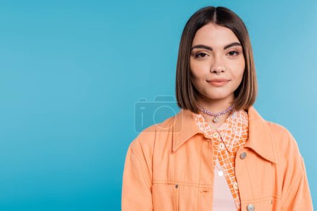 Generation z, Porträt einer hübschen Frau, junges Modemodel, das auf blauem Hintergrund in die Kamera blickt, orangefarbenes Hemd, kurze brünette Haare, durchbohrte Nase, Sommeroutfit, Gen-Z-Mode 