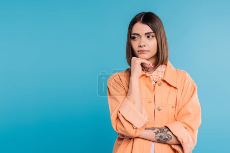 generation z, portrait of concerned woman, young fashion model looking away and thinking on blue background, orange shirt, short brunette hair, pierced nose, summer outfit, gen z fashion 