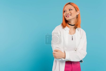 Foto de Felicidad, alegre mujer joven asiática con el pelo teñido de pie en traje casual y sonriendo sobre fondo azul, camisa blanca, mirando hacia otro lado, collar de gargantilla, pelo rojo, generación z - Imagen libre de derechos