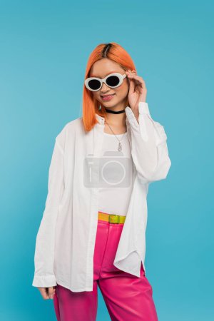 Foto de Joven cultura, alegre mujer asiática con el pelo teñido de pie en traje casual y gafas de sol, sonriendo sobre fondo azul, camisa blanca, collar de gargantilla, pelo rojo, generación z - Imagen libre de derechos