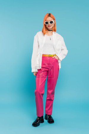happy face, young asian woman with dyed hair standing in casual attire and sunglasses, smiling on vibrant blue background, white shirt, choker necklace, red hair, generation z, hand in pocket