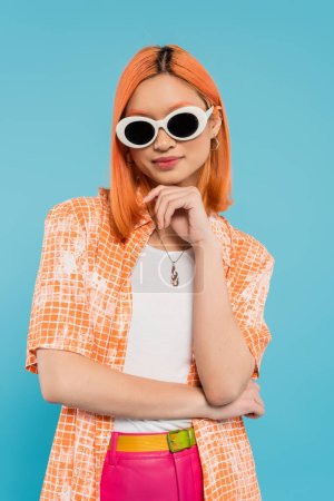 Generation z, junge asiatische Frau mit gefärbten Haaren, lässig gekleidet und Sonnenbrille, Blick in die Kamera auf leuchtend blauem Hintergrund, orangefarbenes Hemd, rote Haare, persönlicher Stil