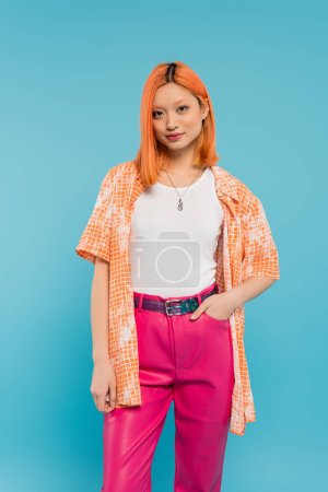 tenue décontractée, jeune femme asiatique aux cheveux rouges teints debout avec la main dans la poche de pantalon rose sur fond bleu vif, chemise orange, style personnel, confiance, génération z 