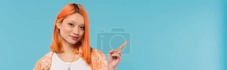 démontrant, jeune femme asiatique avec les cheveux rouges teints en regardant la caméra sur fond bleu vif, chemise orange, pointant du doigt, montrant quelque chose, génération z, bannière 