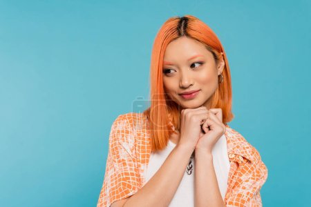 adorable, cheerful young asian woman with dyed red hair smiling and holding hands near face on vibrant blue background, pleased, generation z, casual attire, looking away, young culture 